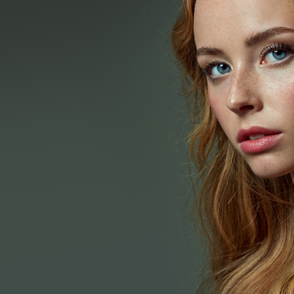 Beautiful young red-haired and fair-skinned girl with freckles posing in the studio on a gray background. Blue eyes.