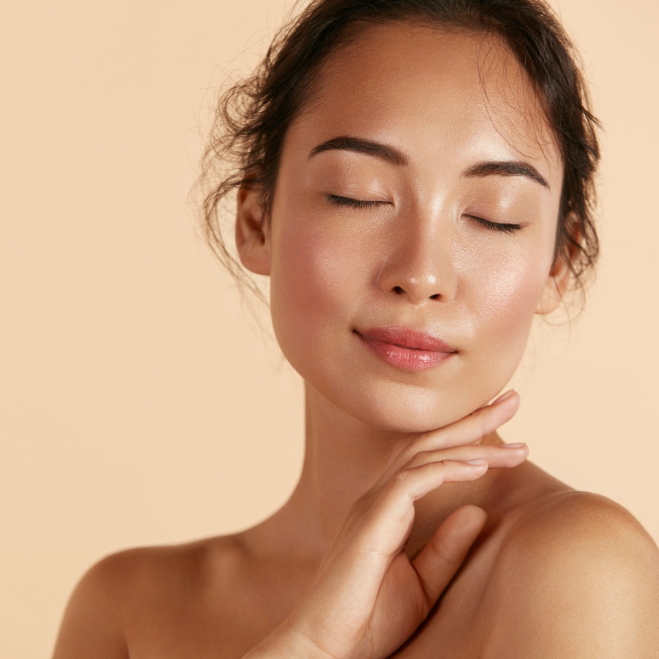 Beauty face. Woman with natural makeup and healthy skin portrait. Beautiful asian girl model touching fresh glowing hydrated facial skin on beige background closeup. Skin care concept