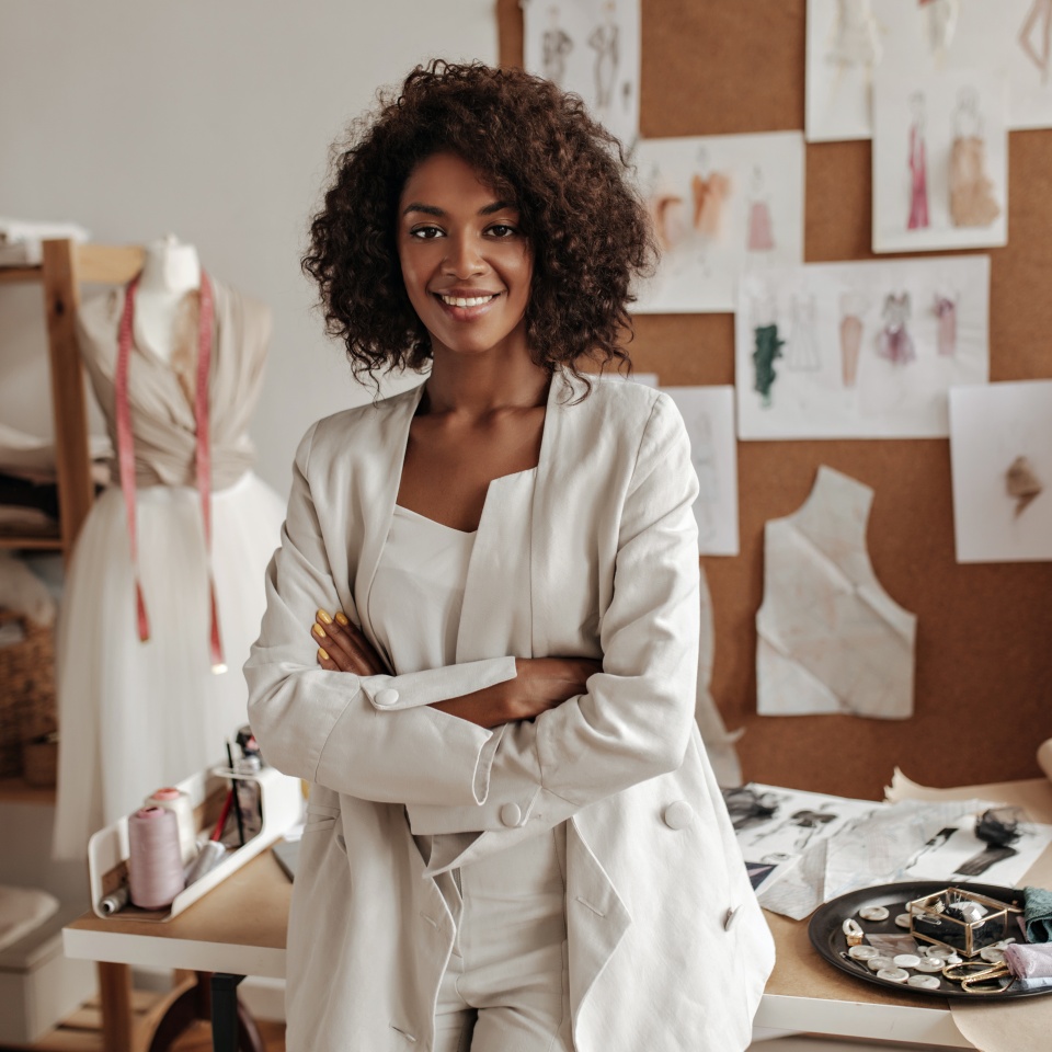 Beautiful curly brunette dark-skinned fashion designer poses in office, leans on table. Young lady in white suit crosses arms and smiles.