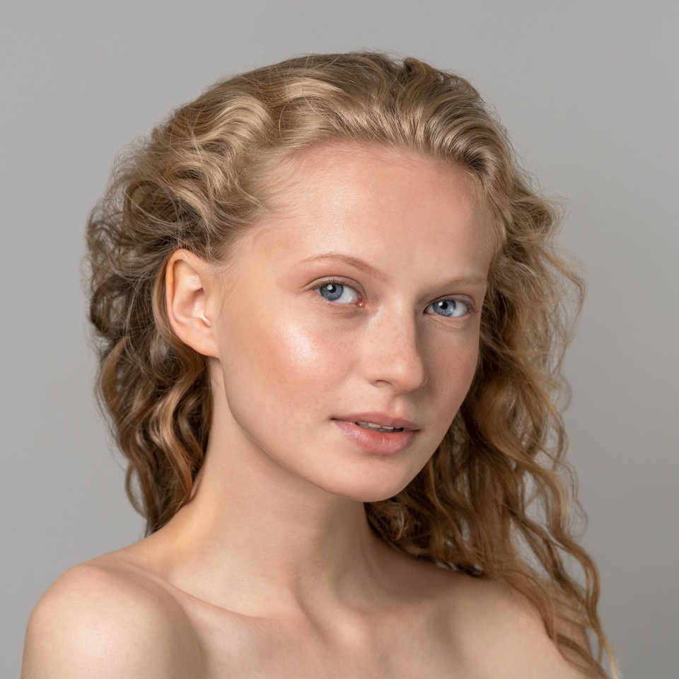 Closeup of pretty young woman face with blue eyes, curly natural blonde hair, has no makeup, touching her soft skin, standing shirtless with bare shoulders, looking at camera. Studio grey background