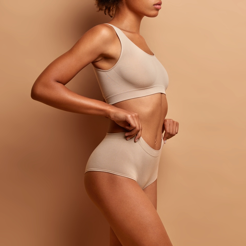 Profile view of slim dark skinned woman dressed in cropped top and panties demonstrates her perfect body slender legs poses against brown background