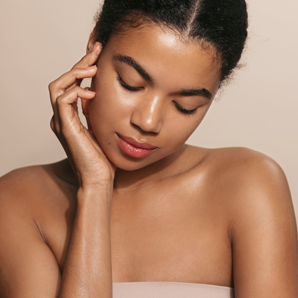 Beautiful african american woman with glowing, nourished head and shoulders, touches her moisturized, perfect facial skin after skincare product, brown background