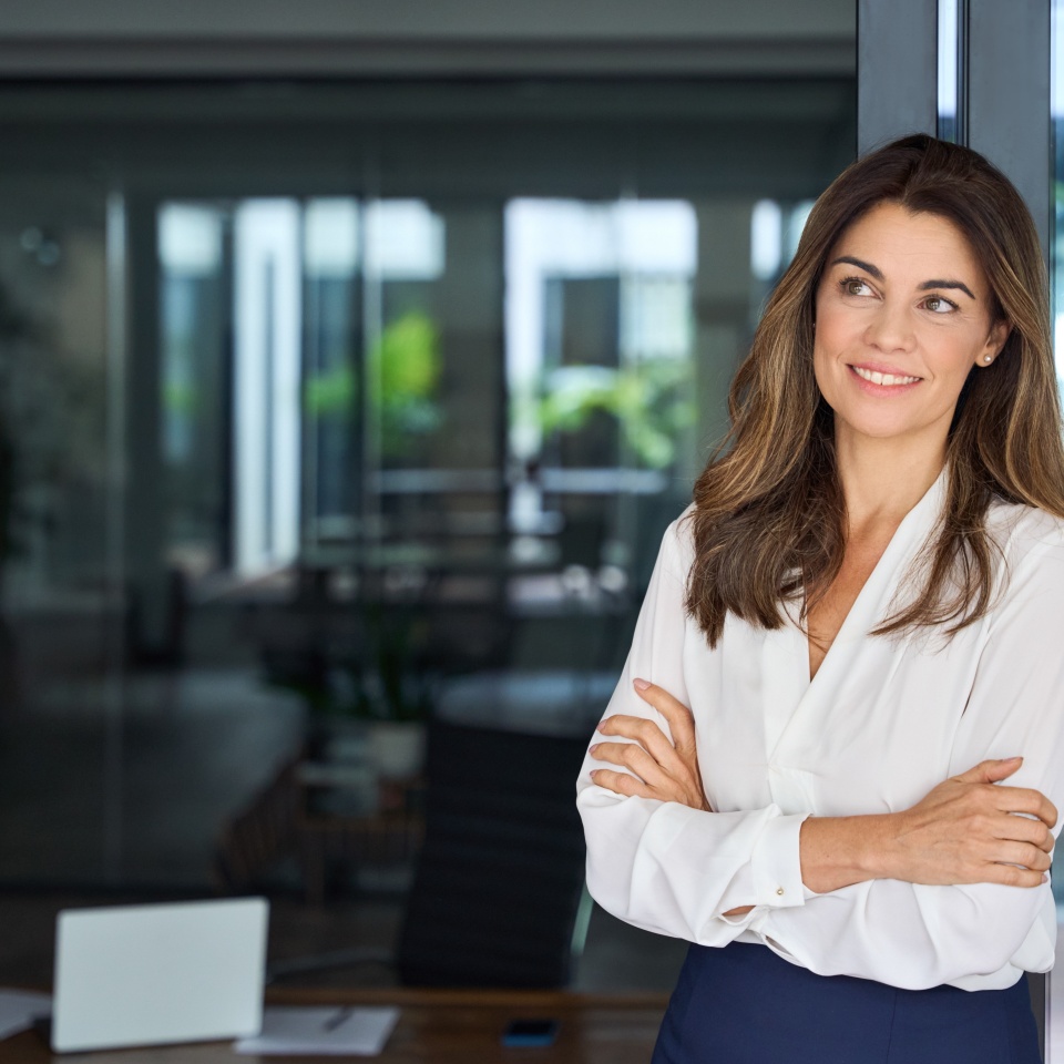 Smiling dreamy Latin professional mid aged business woman corporate manager leader looking away at copy space, happy mature female executive standing at office window arms crossed, thinking.