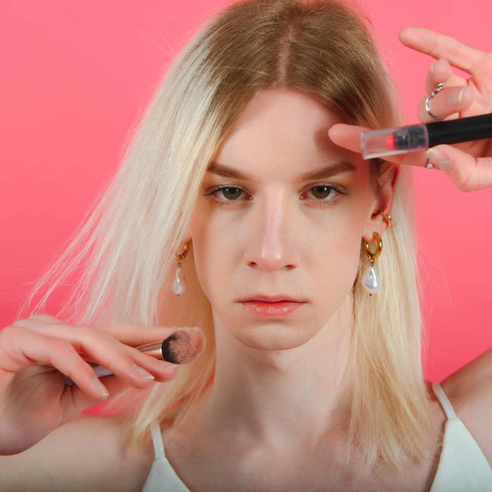 Person with long blonde hair applies makeup with a brush and holds a makeup product, standing against a solid pink background.