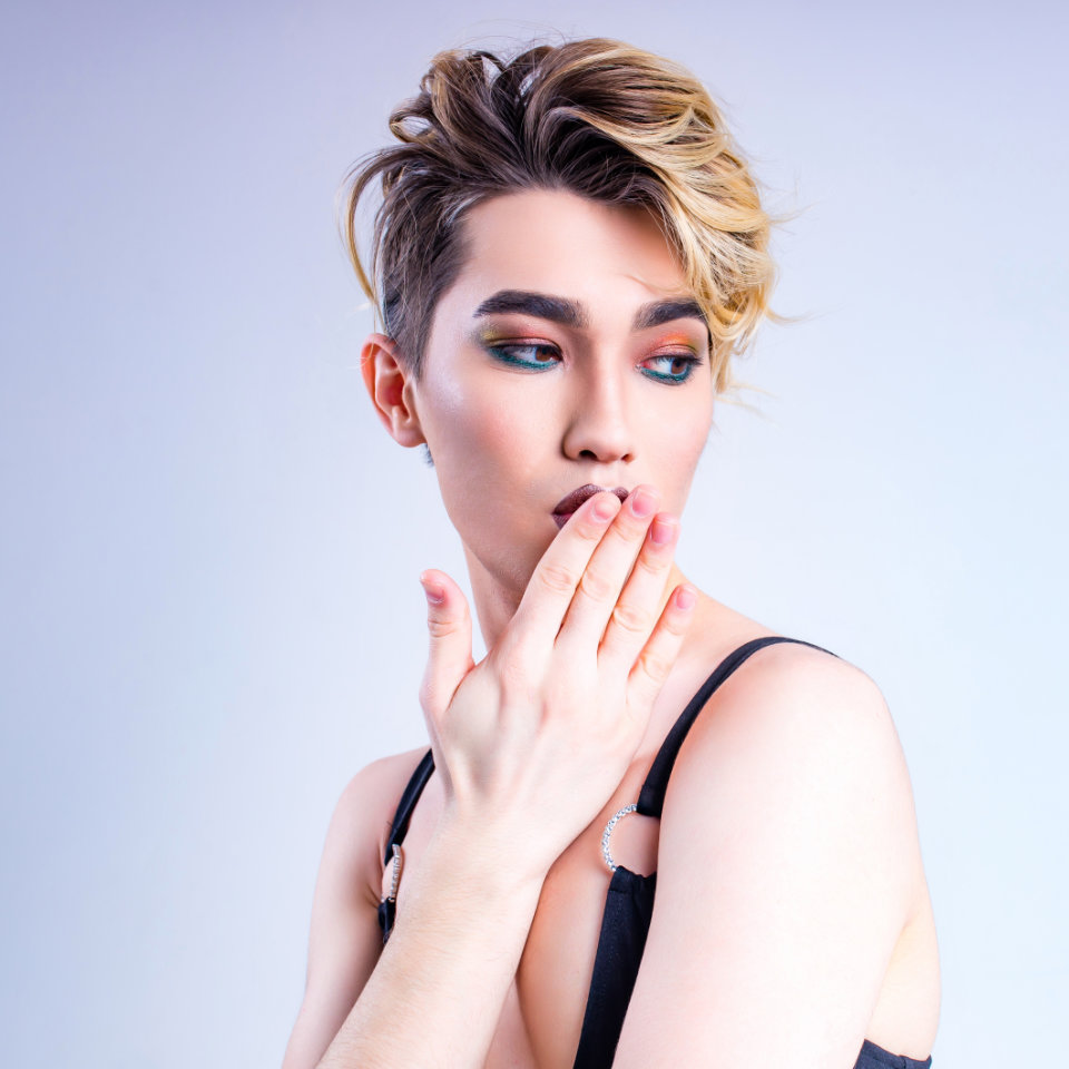 Person with short, styled blond hair and makeup poses against a light background, wearing a black top and touching their lips with their hand.