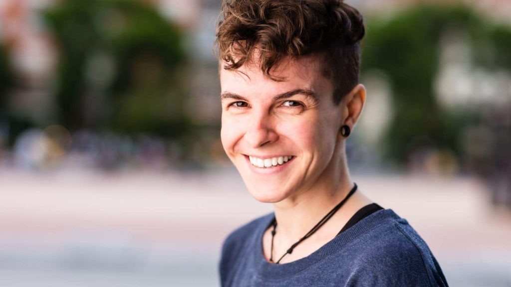 Person with short curly hair and an undercut, wearing a dark shirt and earrings, smiling outdoors with a blurred background of trees and buildings.