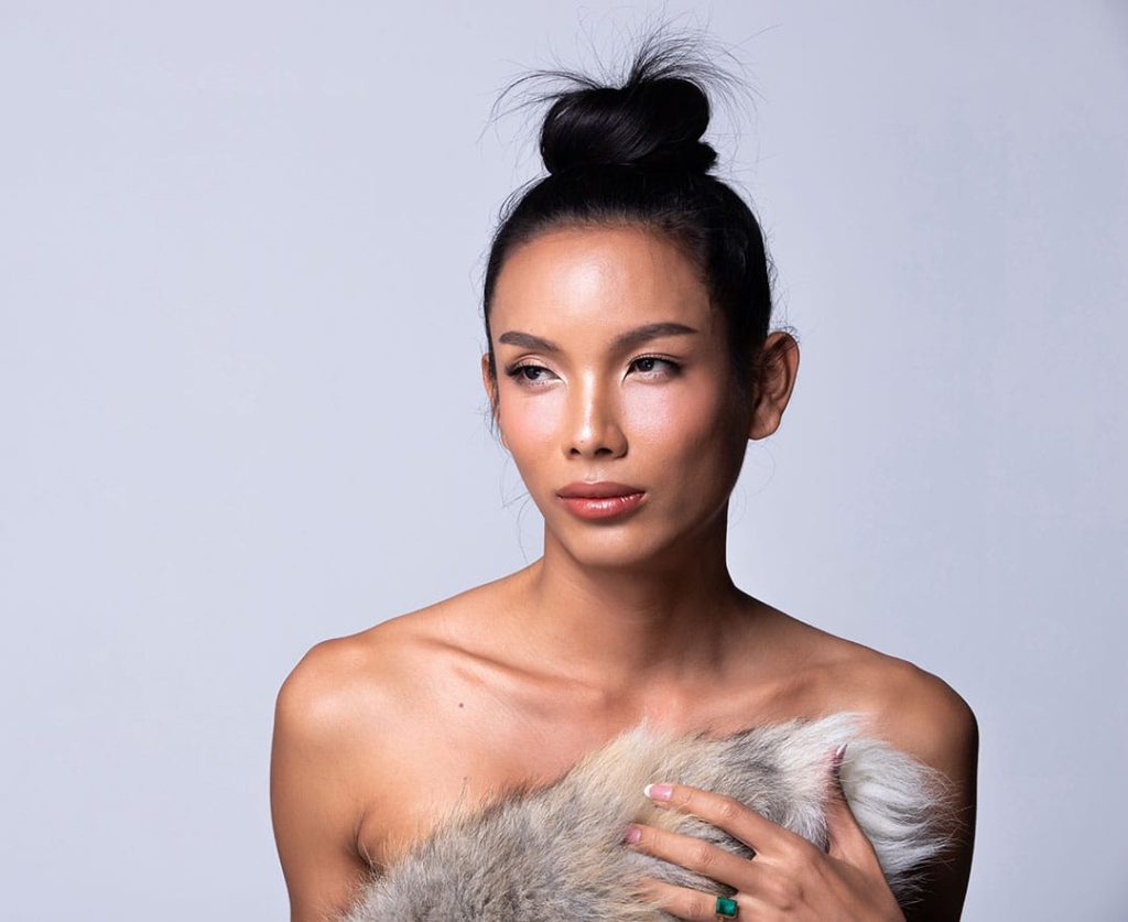 A woman with a topknot hairstyle poses against a plain background, draped in a fur-like fabric, looking slightly to the side with a neutral expression.