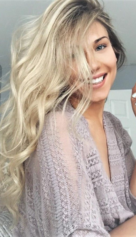 Smiling woman with long, wavy blonde hair wearing a light lavender textured top, standing indoors near a white door.