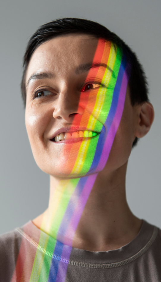 A person with short dark hair smiles as a rainbow-colored light is projected across their face against a plain gray background.
