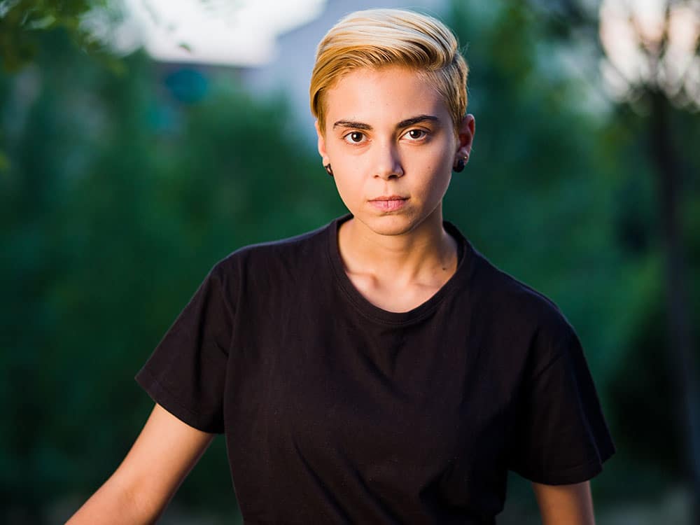 Person with short blond hair wearing a black t-shirt standing outdoors with a neutral expression and green foliage in the background.