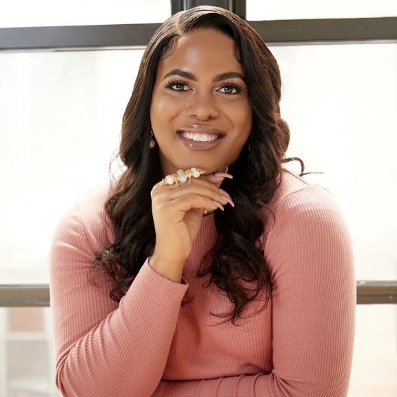 Woman with long wavy hair wearing a pink ribbed sweater sits indoors, smiling and resting her chin on her hand in front of a window.