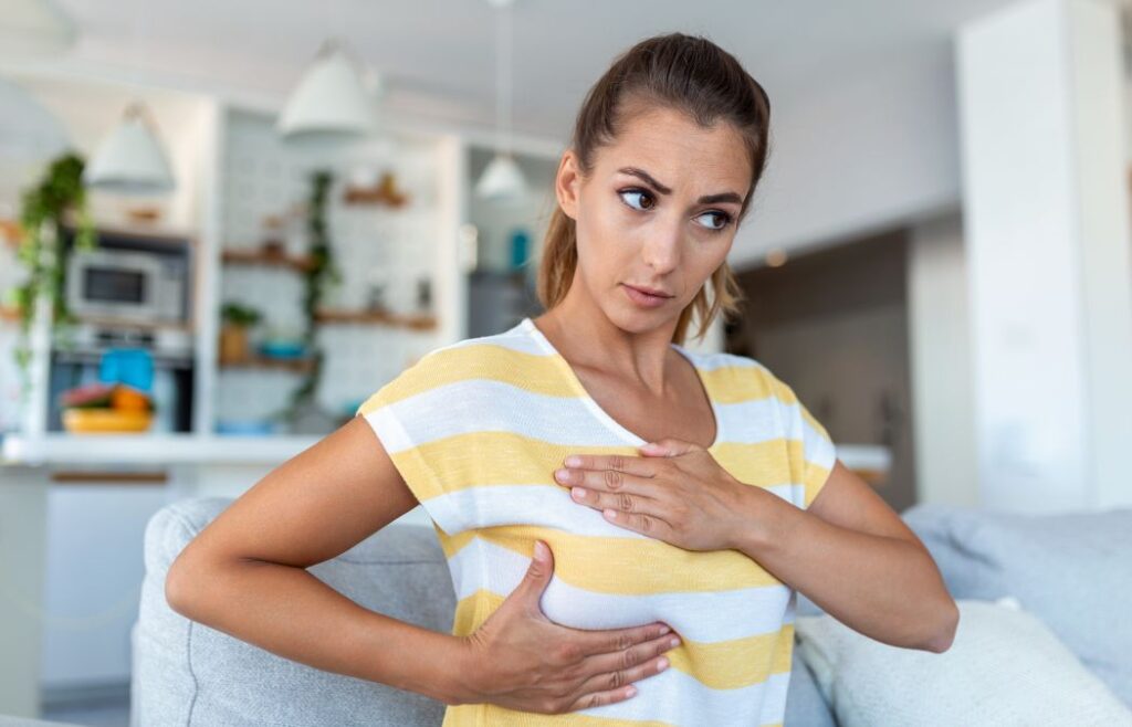 A woman sits on a couch and performs a self-examination of her breast, looking concerned. A kitchen is visible in the background.