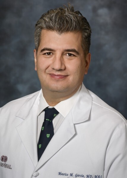 A man with gray hair wearing a white medical coat, dress shirt, and tie, posing in front of a gray studio background.